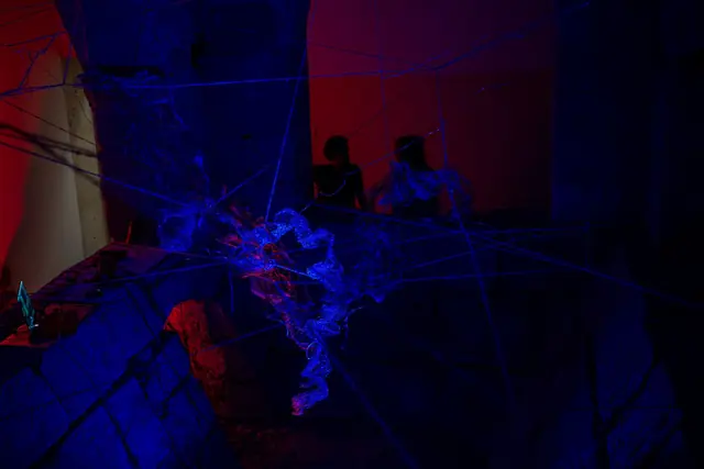 Rhîza: fios de arame iluminados a verde e azul numa sala de pedra com luz vermelha
